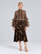 A woman stands against a plain light gray background, wearing a brown ruffled blouse with layered sleeves paired with the Social Queen Floral Beaded Midi Skirt—a brown suede skirt featuring floral embroidery in an elegant A-line silhouette—and gold heels.