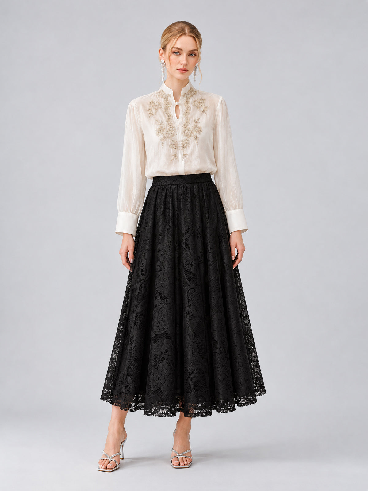 A woman models the Social Queen Floral Vine Embroidered Blouse, featuring delicate floral vine embroidery in ivory, paired with a long black lace skirt and silver heels. She showcases a relaxed straight silhouette against a plain light gray background.