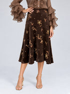 A woman wearing the Social Queen Floral Beaded Midi Skirt, an elegant brown A-line suede midi skirt adorned with floral embroidery, paired with a sheer brown blouse featuring ruffled sleeves and gold strappy heels, stands against a neutral background.