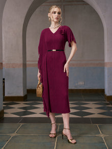 A woman stands indoors wearing the Social Queen Asymmetric Fringe Midi Dress (Belt Included) in burgundy, adorned with delicate 3D floral appliqués. She pairs the dress with gold heels and carries a tan handbag. The scene features tiled floors and arched doorways in the background.