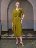 A woman stands indoors on a tiled floor under an archway, wearing the Social Queen Asymmetric Fringe Midi Dress (belt included), paired with gold heels, a necklace, and holding a woven handbag.