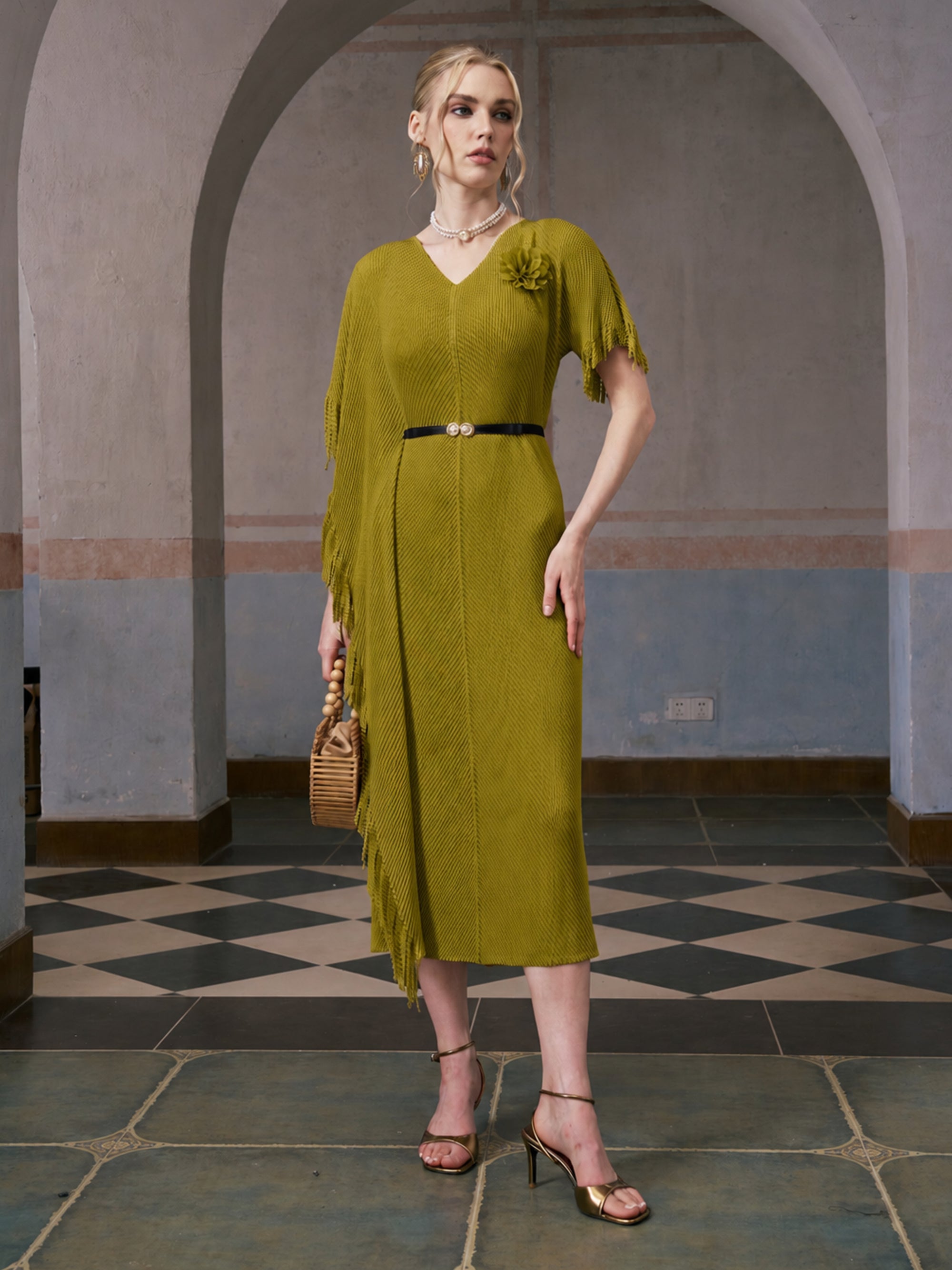 A woman stands indoors on a tiled floor under an archway, wearing the Social Queen Asymmetric Fringe Midi Dress (belt included), paired with gold heels, a necklace, and holding a woven handbag.