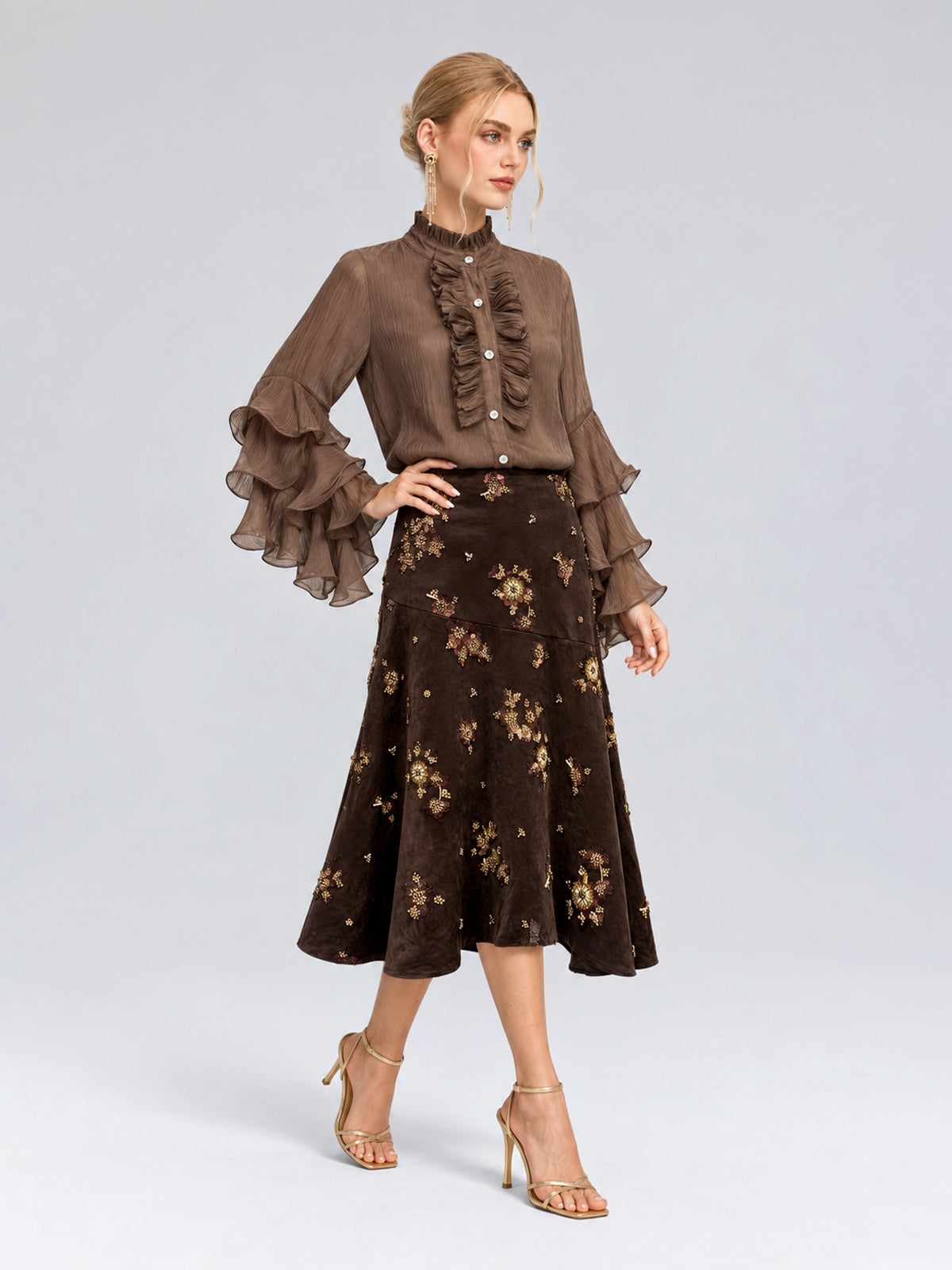 A woman wearing a brown, long-sleeve ruffled blouse and the Social Queen Floral Beaded Midi Skirt, paired with heeled sandals, stands against a plain light background.