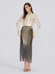 A woman stands against a plain background wearing a cream ruffled blouse, Pearl-Trim Sequin Fringe Skirt by Social Queen, a pearl chain waist belt, and strappy heels.