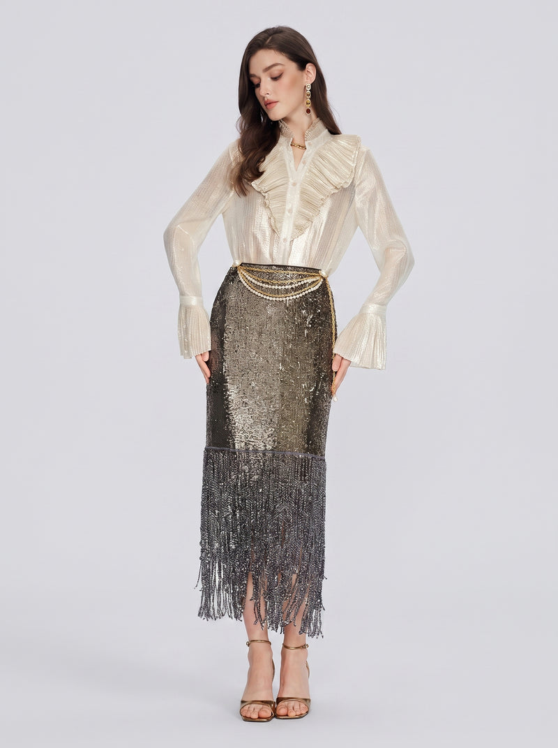 A woman stands against a plain background wearing a cream ruffled blouse, Pearl-Trim Sequin Fringe Skirt by Social Queen, a pearl chain waist belt, and strappy heels.