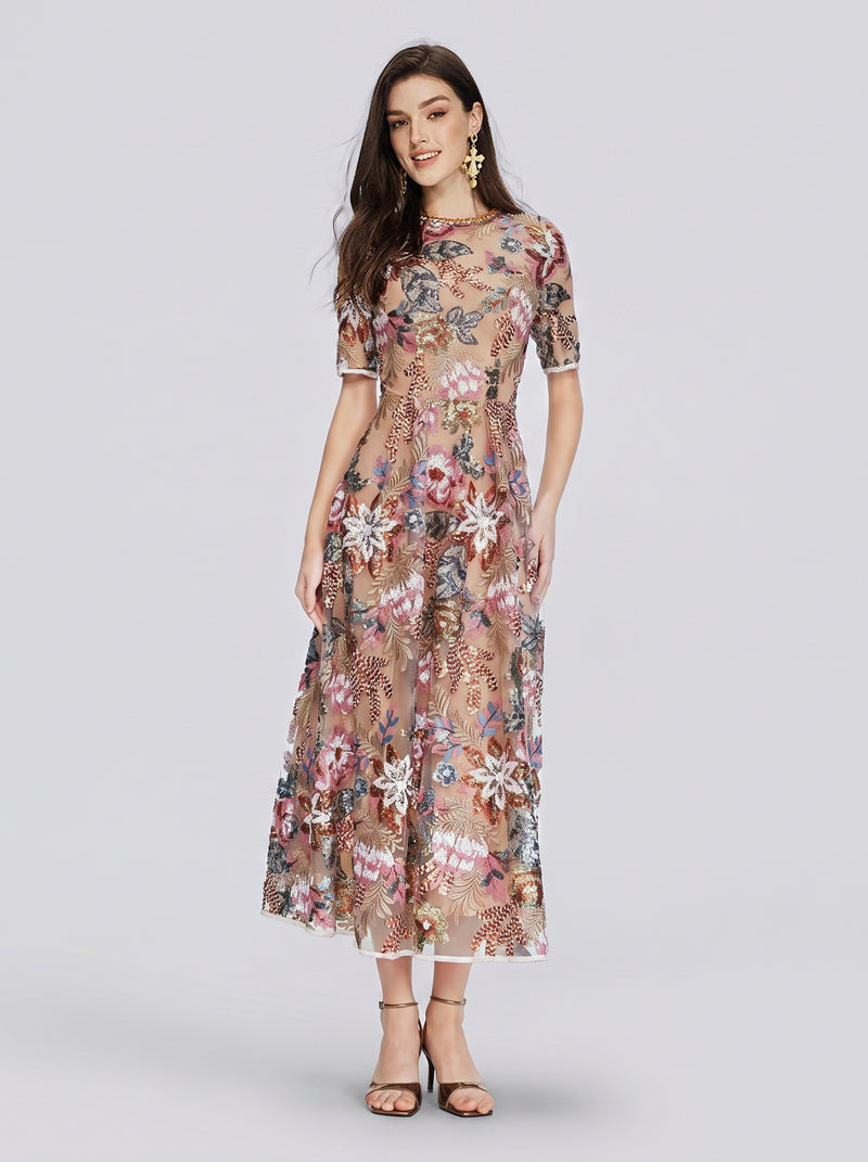 A woman in the Social Queen Sequin Embellished Floral Dress, featuring sheer fabric, floral embroidery, short sleeves, a nude underlay, and paired with black heeled sandals, stands against a plain light background—an elegant ensemble for a wedding guest.