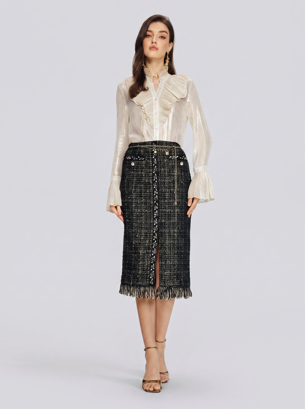 A woman stands facing forward against a plain light gray background, wearing a cream-colored ruffled blouse paired with the Social Queen Hand-Beaded Tweed Fringe Slit Skirt in black and gold, and beige heels.