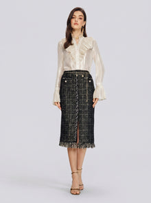 A woman stands facing forward against a plain light gray background, wearing a cream-colored ruffled blouse paired with the Social Queen Hand-Beaded Tweed Fringe Slit Skirt in black and gold, and beige heels.