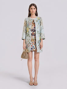A woman stands against a plain light background, wearing the Social Queen Floral Embroidered Silk Jacquard Jacket with its tailored mid-length silhouette over a patterned dress, accessorized with drop earrings, sandals, and a small woven bag.