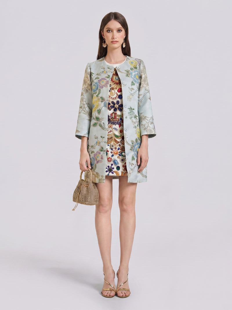 A woman stands against a plain light background, wearing the Social Queen Floral Embroidered Silk Jacquard Jacket with its tailored mid-length silhouette over a patterned dress, accessorized with drop earrings, sandals, and a small woven bag.