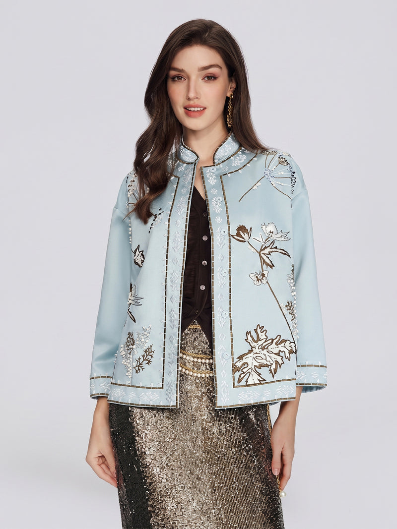 A woman stands facing forward against a plain light background, wearing the Social Queen Floral Embroidered Beaded Jacket—a light blue, stand-collar silk piece embellished with hand-embroidered floral motifs—over a black top and a gold sequin skirt.