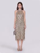 A woman wearing the Social Queen Crystal-Embellished Sequin Fringe Halter Dress—sleeveless, knee-length, and adorned with metallic details—holds a small round purse and pairs the look with heeled sandals while standing against a plain light background.