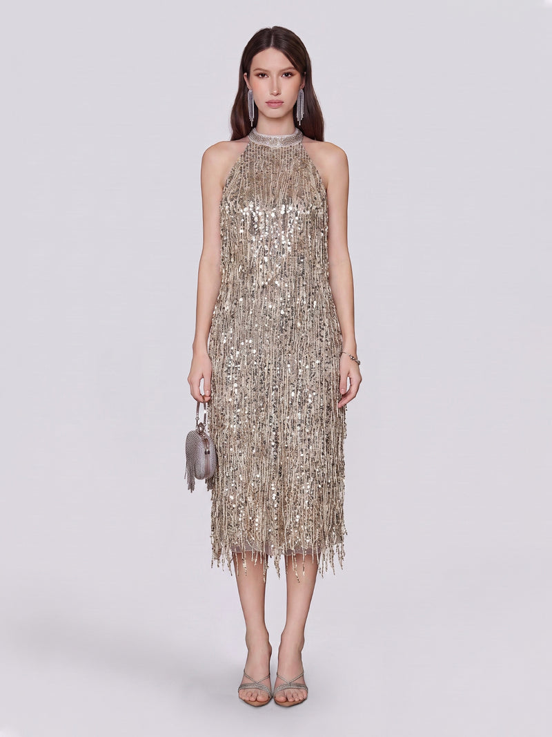 A woman wearing the Social Queen Crystal-Embellished Sequin Fringe Halter Dress—sleeveless, knee-length, and adorned with metallic details—holds a small round purse and pairs the look with heeled sandals while standing against a plain light background.