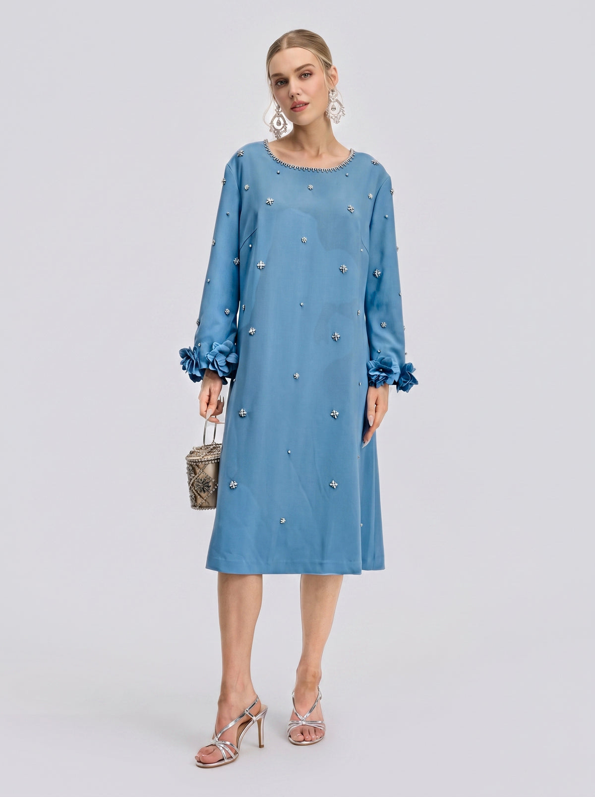 A woman stands wearing the Social Queen Handcrafted Beaded Floral A-Line Dress in light blue, featuring a knee-length silhouette with long sleeves and intricate hand-beaded floral appliqués and embellishments; she accessorizes with a small bag and silver sandals.