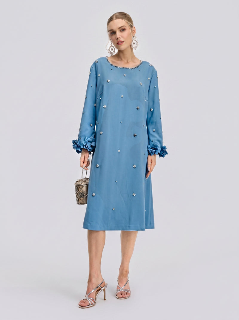 A woman stands wearing the Social Queen Handcrafted Beaded Floral A-Line Dress in light blue, featuring a knee-length silhouette with long sleeves and intricate hand-beaded floral appliqués and embellishments; she accessorizes with a small bag and silver sandals.
