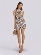A woman wearing the Social Queen Jewel-Embellished Floral Mini Dress, styled with sandals and holding a small woven handbag, stands against a plain light background.