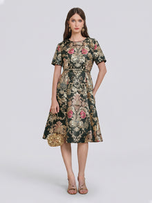 A woman stands against a plain light background wearing the Social Queen 100% Silk Jacquard Satin Gold Embroidered Floral Dress, a knee-length piece featuring short sleeves, pink details, and luxurious gold floral embroidery. She accessorizes with a gold clutch and strappy sandals.