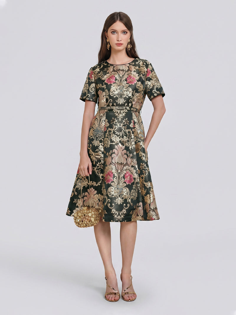 A woman stands against a plain light background wearing the Social Queen 100% Silk Jacquard Satin Gold Embroidered Floral Dress, a knee-length piece featuring short sleeves, pink details, and luxurious gold floral embroidery. She accessorizes with a gold clutch and strappy sandals.