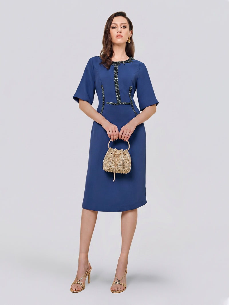 A woman stands against a plain light background wearing the Social Queen Elegant Hand-Beaded Green Crystal Dress—a knee-length blue midi dress with short sleeves, delicately embellished with hand-sewn green crystals—paired with heels and a woven handbag.