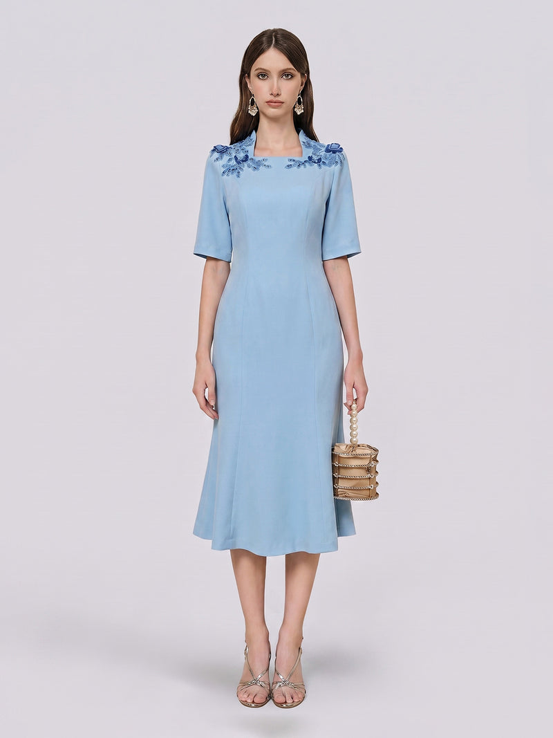 A woman stands against a plain light background wearing the Social Queen Handcrafted Beaded Floral Dress—a sky-blue knee-length design adorned with delicate 3D floral appliqués and blue beaded details. She pairs the look with silver heels and a woven handbag.