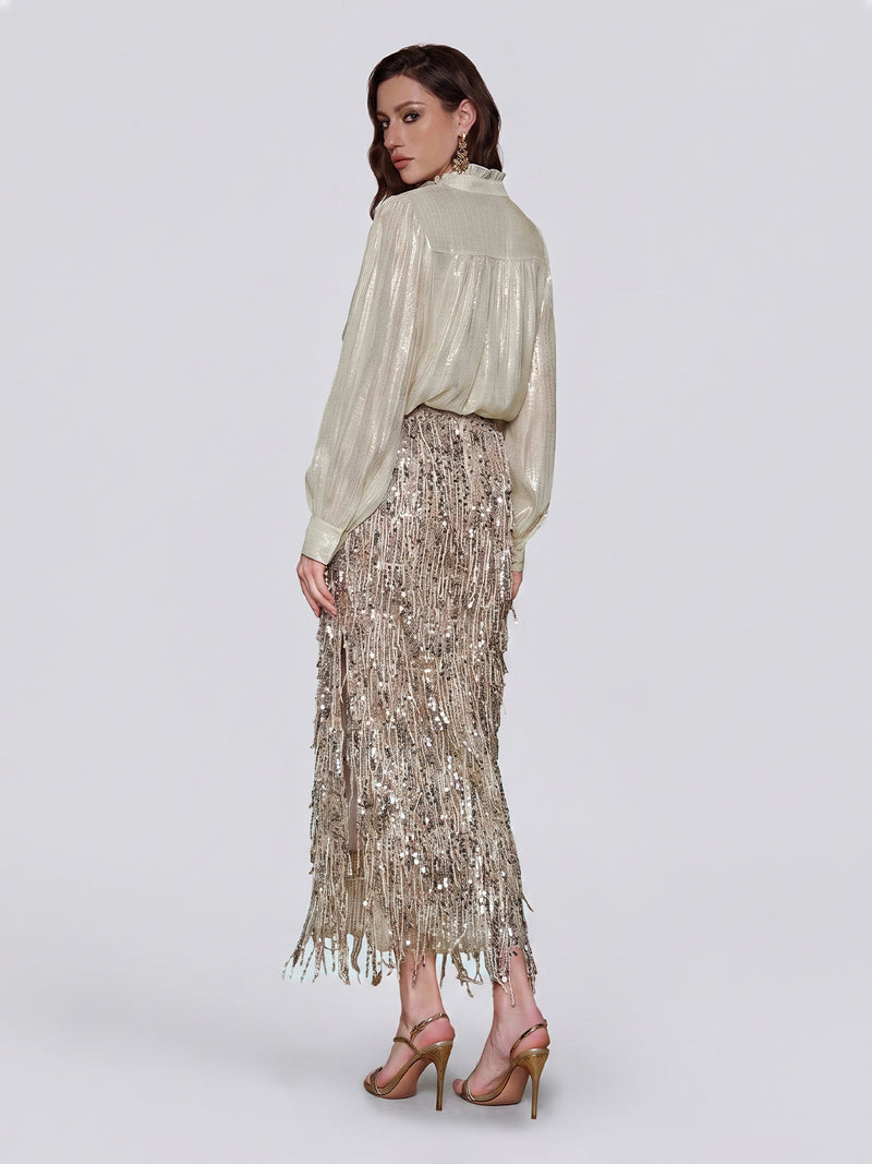 A woman stands in profile against a plain light background, wearing the Social Queen Sequin Fringe Side-Slit Midi Skirt styled with a shimmery long-sleeve blouse tucked in and high-heeled sandals.