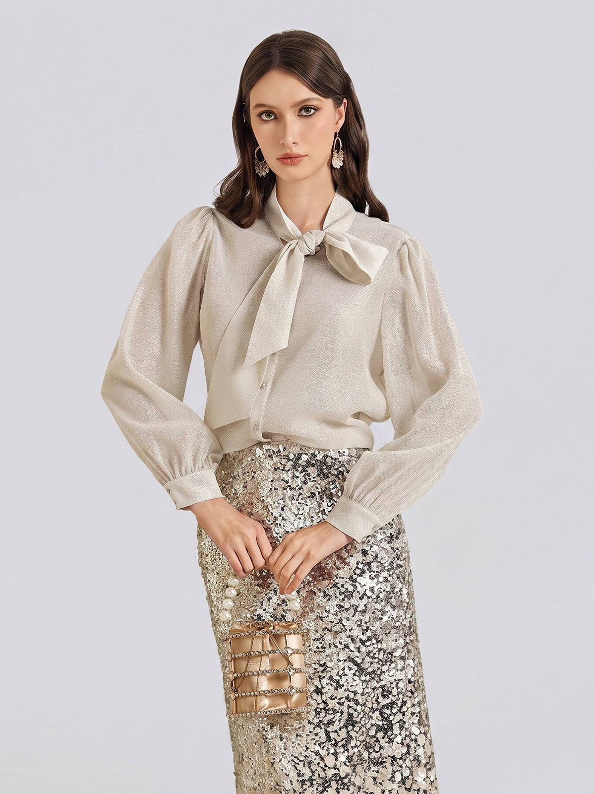 A woman stands against a plain light background wearing a cream blouse with a bow and long sleeves, paired with the Social Queen Ombre Silver & Gold Sequin Slim Skirt, and holding a small metallic bag.