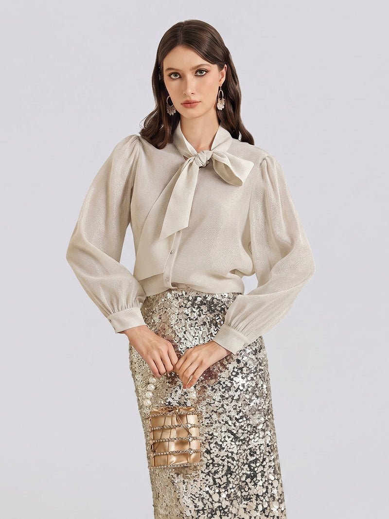 A woman stands against a plain light background wearing a cream blouse with a bow and long sleeves, paired with the Social Queen Ombre Silver & Gold Sequin Slim Skirt, and holding a small metallic bag.