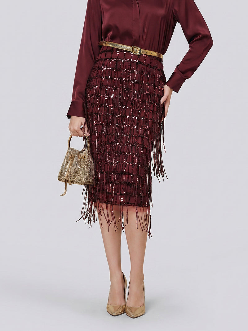 A person is wearing the Social Queen Hand-Embellished Sequin Fringe Skirt, paired with a burgundy blouse, tan belt, gold heels, and a gold purse, as they stand against a plain gray background.