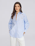 A woman stands and smiles against a plain light gray background, wearing the Social Queen Pearl & Beaded Striped Blouse in light blue, styled with white pants.