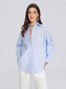 A woman stands and smiles against a plain light gray background, wearing the Social Queen Pearl & Beaded Striped Blouse in light blue, styled with white pants.