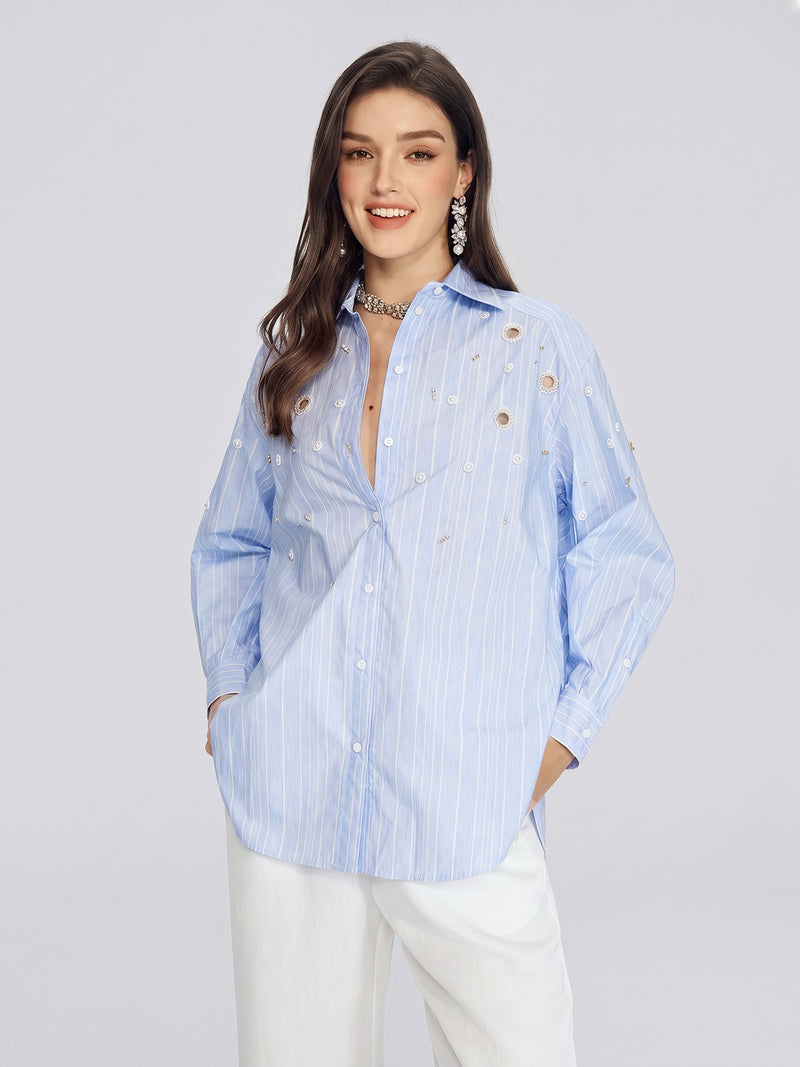 A woman stands and smiles against a plain light gray background, wearing the Social Queen Pearl & Beaded Striped Blouse in light blue, styled with white pants.