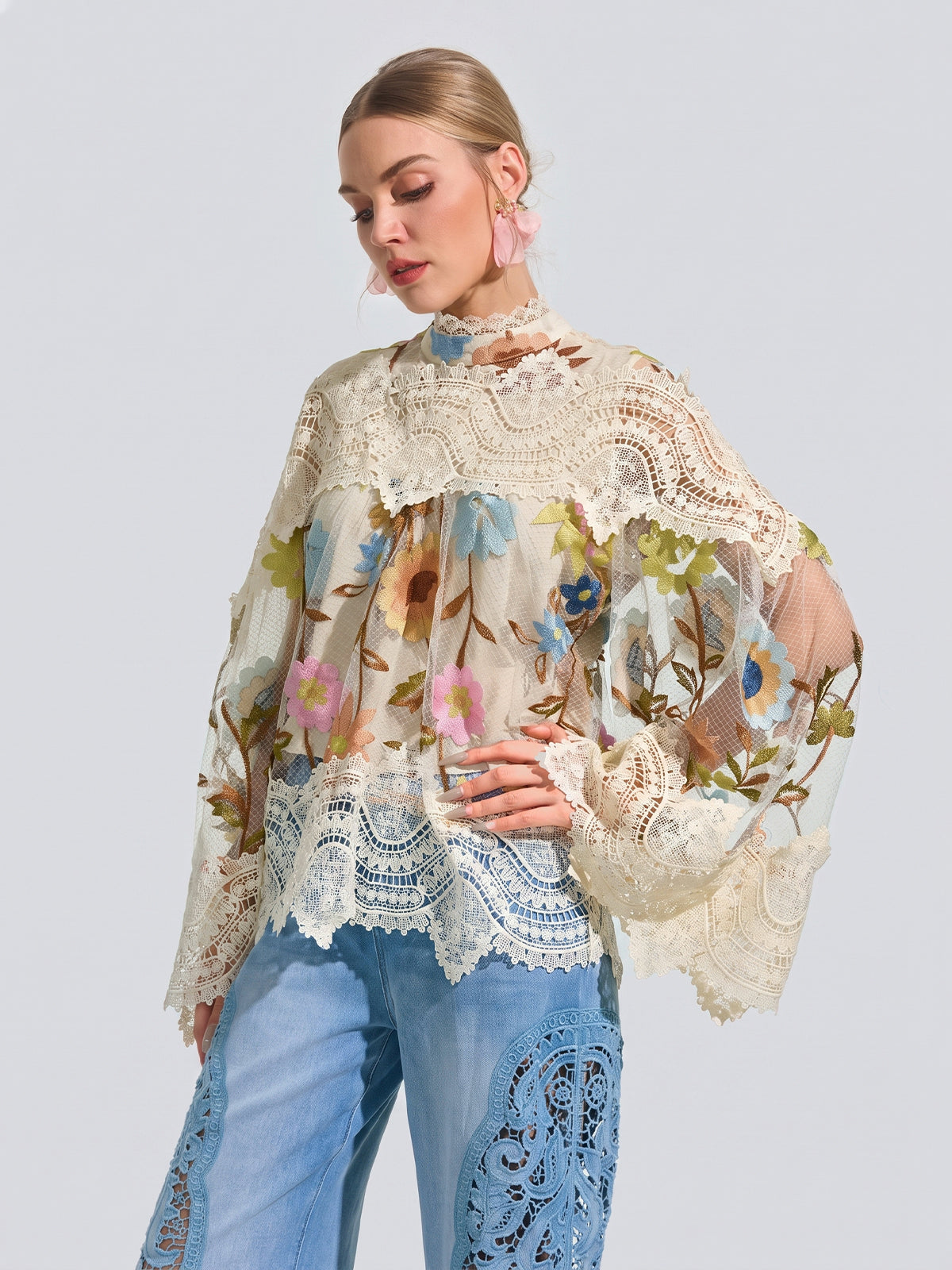A woman wears the Social Queen Floral Embroidered Lace Trim Top—featuring sheer, long sleeves, a high neck, and delicate floral embroidery—paired with light blue jeans with lace detailing, standing against a light gray background.