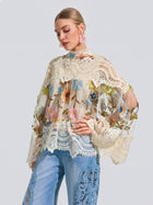 A woman wears the Social Queen Floral Embroidered Lace Trim Top—featuring sheer, long sleeves, a high neck, and delicate floral embroidery—paired with light blue jeans with lace detailing, standing against a light gray background.