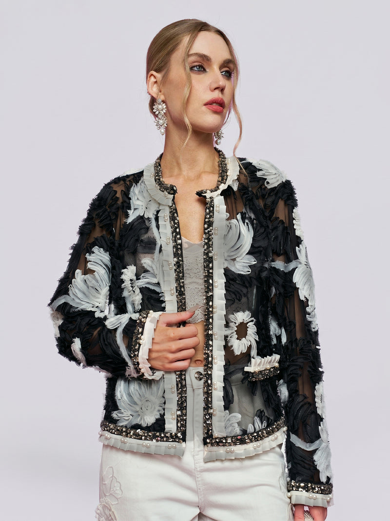 A woman stands against a plain light background wearing the Social Queen Floral Embroidered Tulle Beaded Jacket, featuring delicate black and white 3D floral embroidery, paired with white pants and large statement earrings.