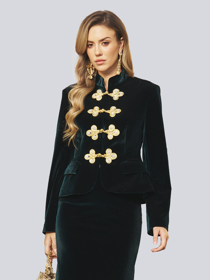 A woman wears the Social Queen Velvet Knot-Button Jacket—a black velvet holiday party blazer with ornate gold buttons—paired with the matching skirt and a gold handbag, standing against a plain light background.