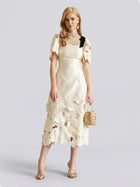 A woman stands against a plain light gray background, wearing the Social Queen Cutout Floral Appliqué Satin Dress in ivory with short sleeves, styled with gold heels and a beige handbag.