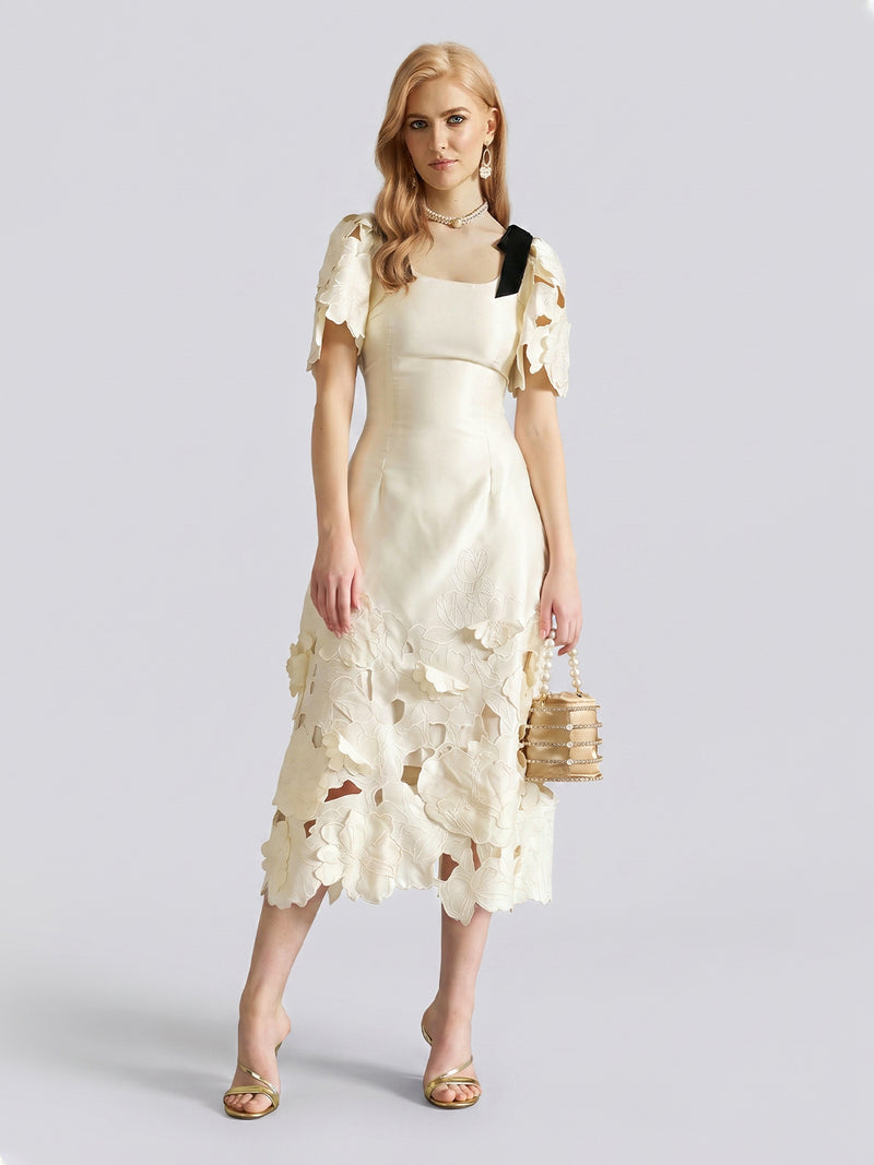 A woman stands against a plain light gray background, wearing the Social Queen Cutout Floral Appliqué Satin Dress in ivory with short sleeves, styled with gold heels and a beige handbag.