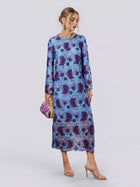 A woman wearing the Social Queen Crystal Trim Lace Shift Dress, featuring long sleeves in dimensional blue and purple lace with delicate floral details and a crystal-embellished neckline, holds a small clutch and stands in high-heeled sandals against a plain background.