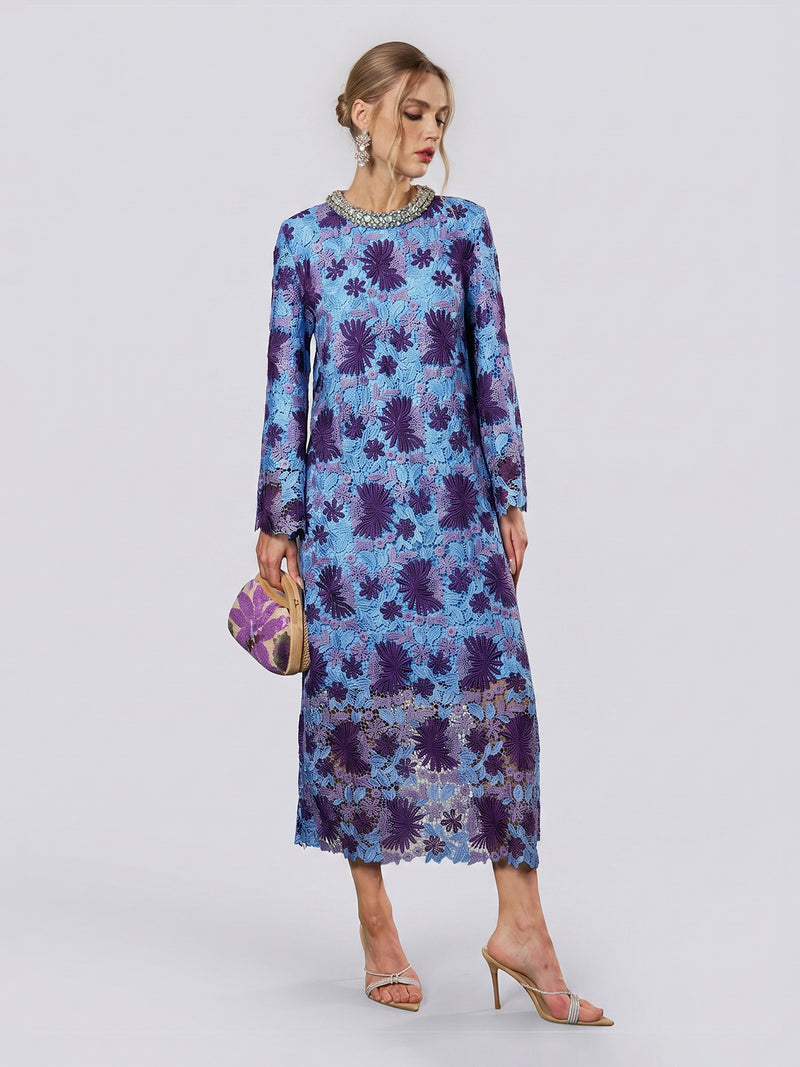 A woman wearing the Social Queen Crystal Trim Lace Shift Dress, featuring long sleeves in dimensional blue and purple lace with delicate floral details and a crystal-embellished neckline, holds a small clutch and stands in high-heeled sandals against a plain background.
