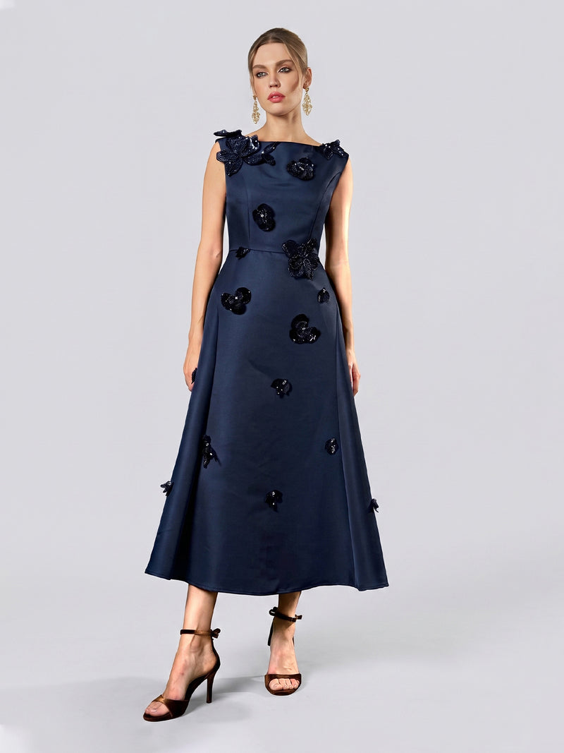 A woman stands against a plain light gray background wearing the Social Queen Floral Appliqué Satin Dress, a sleeveless, navy blue, ankle-length satin gown featuring delicate floral appliqué. She completes her look with black high heels and hoop earrings—an elegant choice for formal occasions.