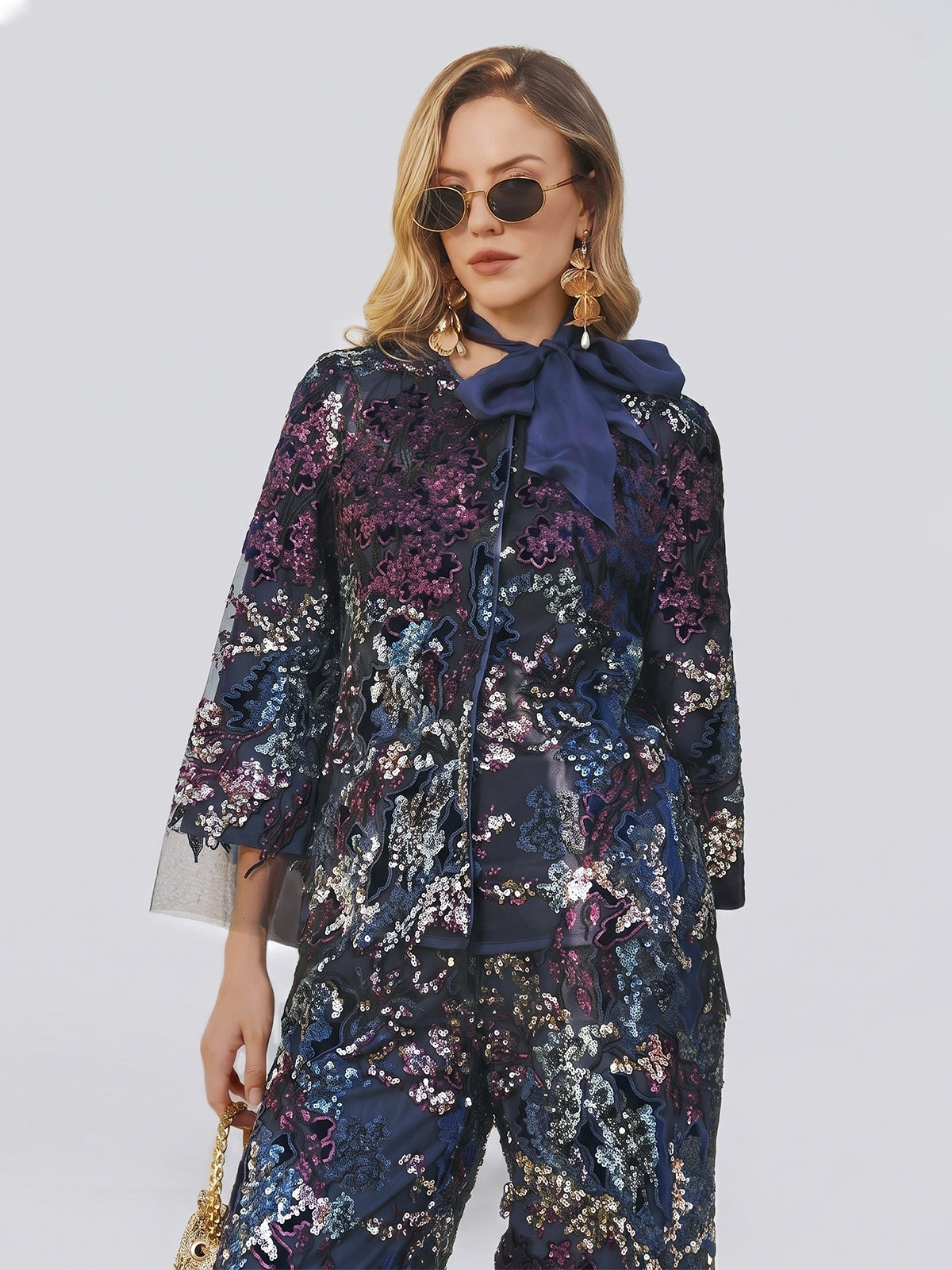 A woman wears the Social Queen Sequin Embroidered Velvet Mesh Jacket styled as a chic pantsuit—featuring sheer sleeves, a large navy bow at the collar, and navy floral detailing. She accessorizes with statement drop earrings, sunglasses, and a gold handbag for a modern wedding guest look against a plain background.