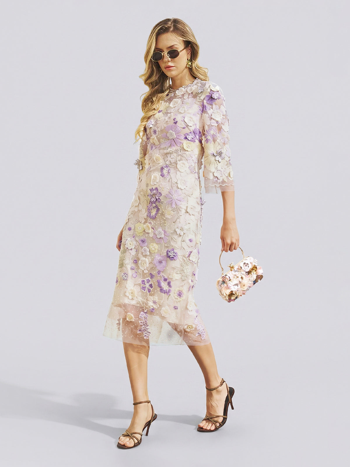 A woman in sunglasses wears the Social Queen 3D Floral Embroidered Beaded Dress—a sheer, knee-length design with purple and white floral appliqués and delicate beadwork—paired with a matching floral handbag and black strappy heels as she walks in front of a plain background. The dress’s subtle cheongsam-inspired silhouette offers an elegant, fashion-forward option for any stylish wedding guest.