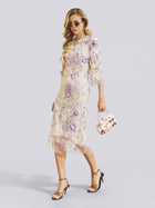 A woman in sunglasses wears the Social Queen 3D Floral Embroidered Beaded Dress—a sheer, knee-length design with purple and white floral appliqués and delicate beadwork—paired with a matching floral handbag and black strappy heels as she walks in front of a plain background. The dress’s subtle cheongsam-inspired silhouette offers an elegant, fashion-forward option for any stylish wedding guest.