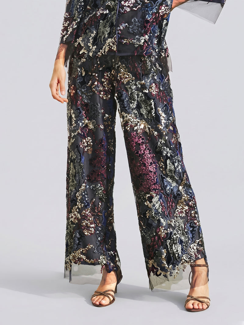 A person is wearing the Sequin Embroidered Mesh Wide-Leg Pants by Social Queen—high-waisted, sheer black pants adorned with multicolored sequin embroidery—paired with open-toe sandals; upper body partially visible, creating a chic look ideal for a stylish wedding guest.