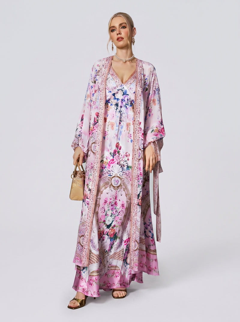 A woman stands against a plain white background, wearing Social Queen’s Baroque Print Mulberry Silk Robe with Scarf draped over a long, flowing floral pink and white dress, and holding a small tan handbag.