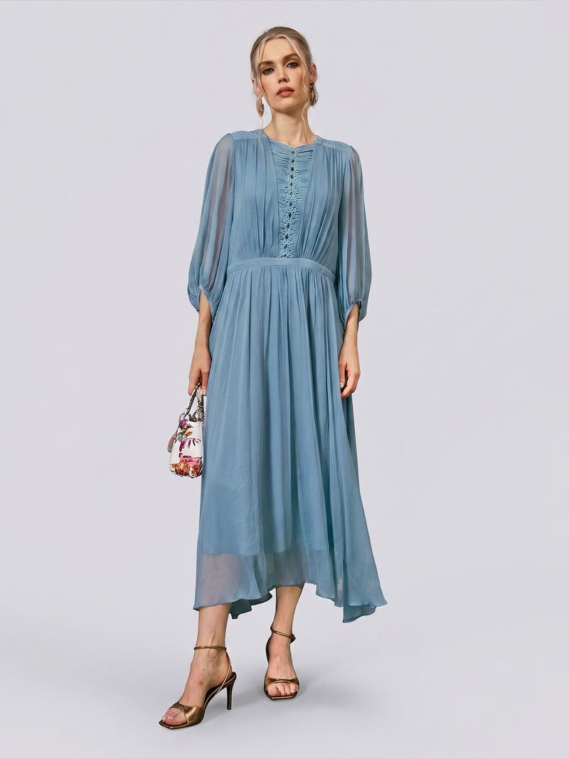 A woman wearing the Social Queen Woven Cutwork Silk Dress—a light blue, long-sleeved, flowy mulberry silk dress with an A-line silhouette—pairs it with gold heels and holds a small floral handbag, standing against a plain light background.