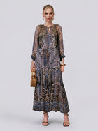 A woman stands against a plain background wearing the Social Queen Leopard Paisley Print Silk Dress, featuring full-length mulberry silk with subtle beaded rhinestone details. She completes the look with a wicker handbag, heeled sandals, and gold jewelry.