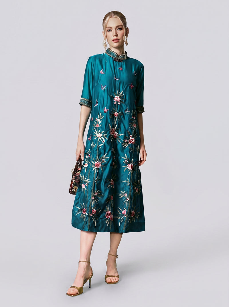 A woman wearing the Social Queen Botanical Embroidered Tencel Dress in peacock green, styled with gold sandals and a small handbag, stands against a plain light background.