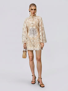 A woman stands against a plain background wearing the Social Queen Baroque Print Floral Appliqué Dress, paired with black heels and a small beige handbag. Her hair is styled up, and she accessorizes with large hoop earrings.