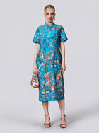 A woman stands facing forward wearing the Social Queen Allover Bird-Embroidered Dress, a turquoise, knee-length piece adorned with colorful bird and floral patterns. She holds a small handbag and wears strappy sandals.